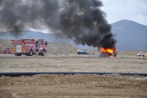 A car on fire with billowing black smoke in a dirt field and with a firetruck standing.